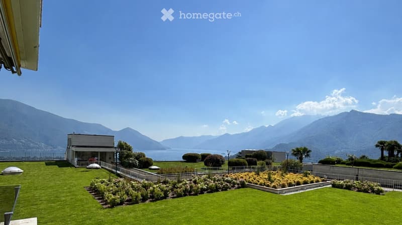 Well-maintained garden with neatly trimmed hedges, flower beds, and a small building, overlooking a scenic lake and mountain range.
