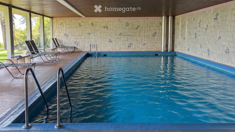 Indoor swimming pool area with lounge chairs, tiled walls, and large windows overlooking a green outdoor space.