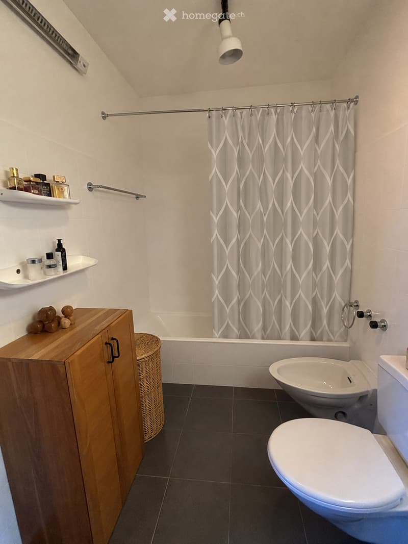 Modern bathroom with dark tiled floor, white walls, wooden cabinet, wicker basket, shelves with toiletries, bathtub, toilet, and shower curtain.