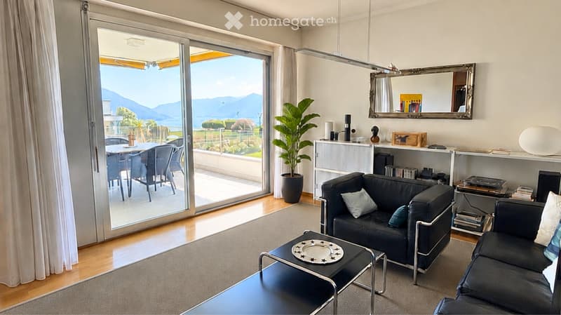 Modern living room with large windows and a view, featuring a black leather sofa, coffee table, dining area, and decorative items.