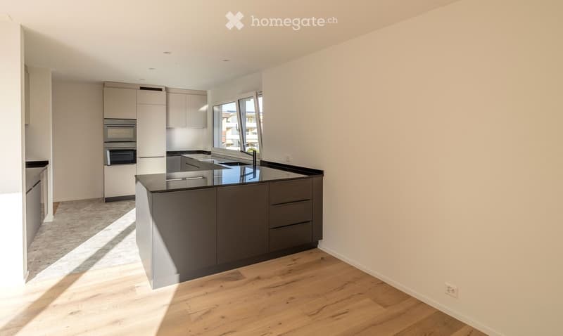 Modern kitchen with a central island, stainless steel sink, and built-in ovens. Light wood flooring and white walls.