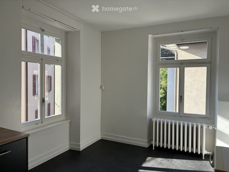 Empty room with light grey walls, dark flooring, and two large windows with white frames. A white radiator is placed beneath one window, and a small wooden cabinet is visible on the left side.