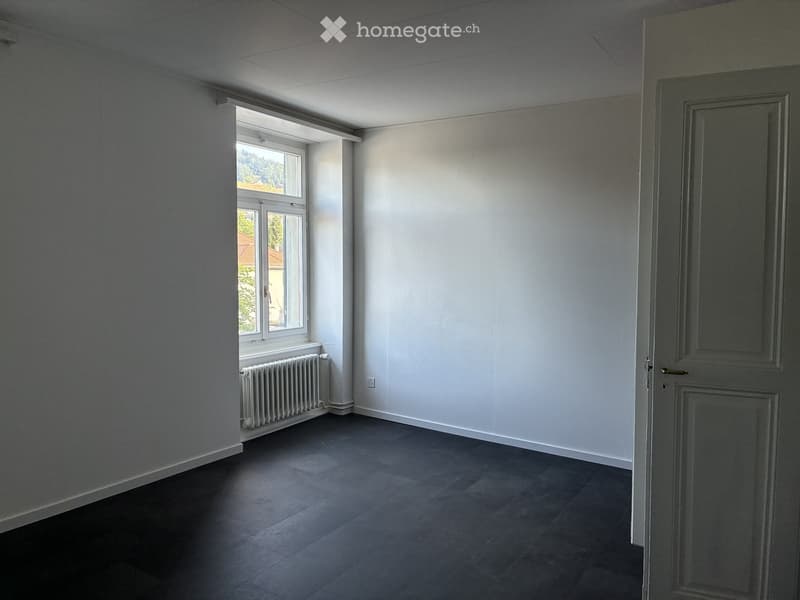 Empty room with white walls, dark tiled floor, and a window with a radiator below it. The room has a door on the right side.