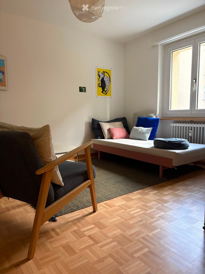 A modern, minimalist bedroom with a wooden floor, a bed with multiple pillows, a sofa, a small round table, and artwork on the walls.