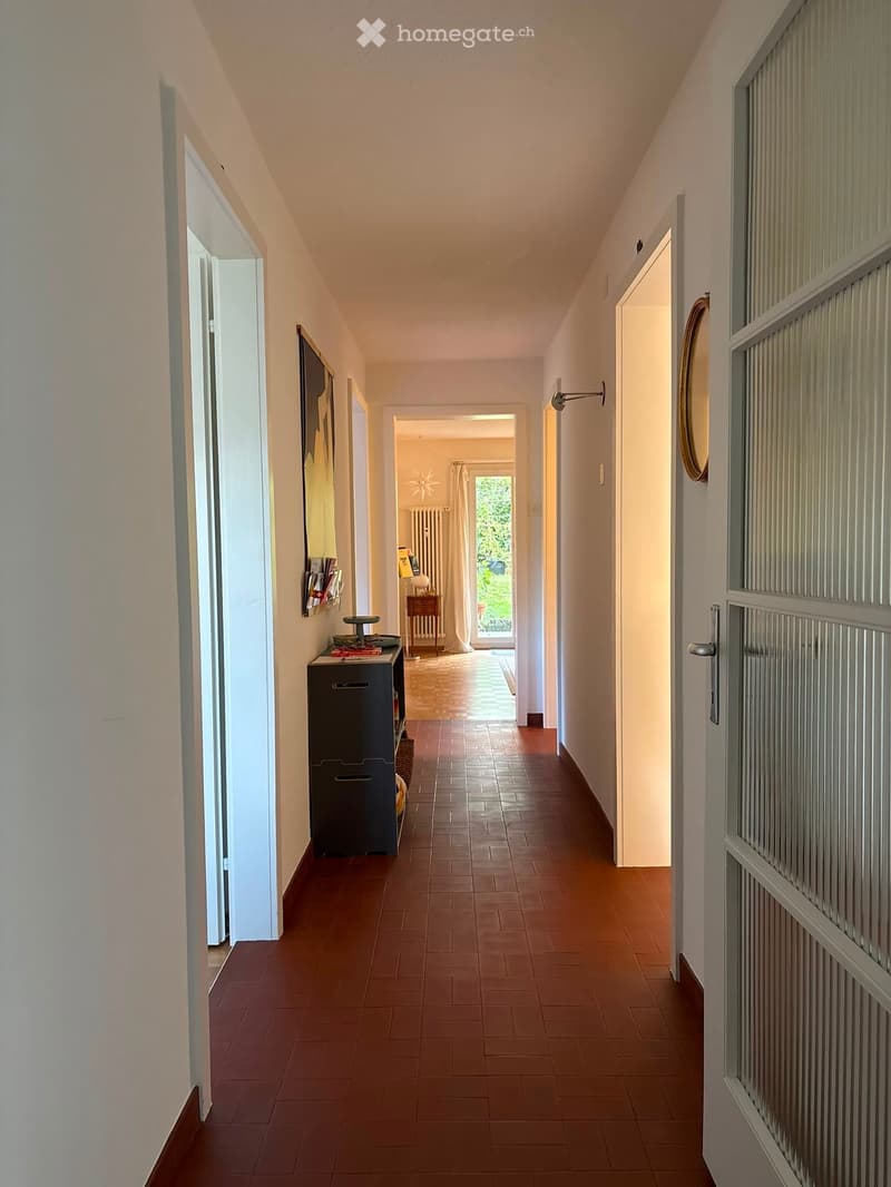 A long, narrow hallway with terracotta tiled flooring, light-colored walls, and several doors leading to other rooms. A small table with books and a mirror is visible on the left side.
