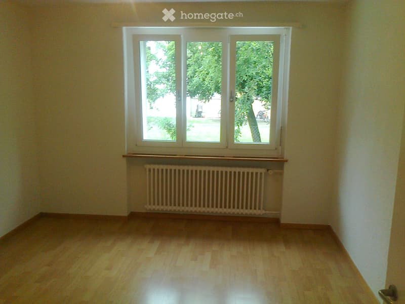 Empty room with wooden flooring, large windows, radiator, and a small wooden shelf above it.