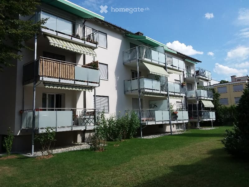 Modern multi-story apartment building with balconies, green roof, and surrounding lawn.