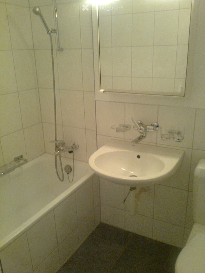 Small bathroom with a bathtub, sink, and showerhead. Light-colored tiles cover the walls and floor.