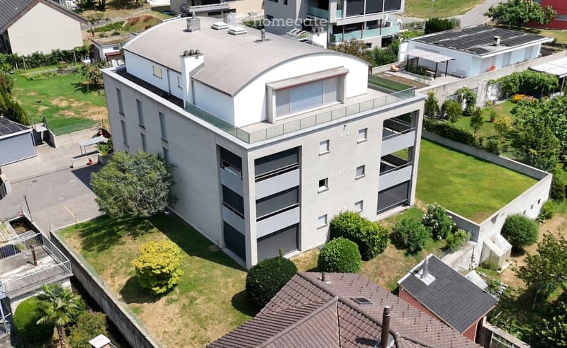 Aerial view of a modern, two-story house with a flat roof, large windows, and a balcony on the upper level. The house is surrounded by a well-maintained garden with trees and shrubs, and there are neighboring houses and solar panels visible in the backgro
