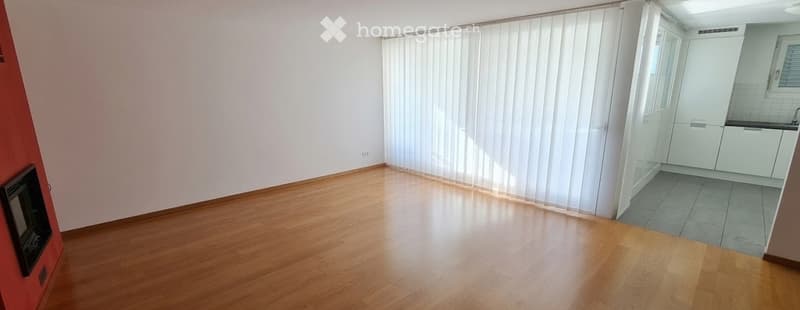 Empty living room with wooden flooring, white walls, and a small kitchen area visible in the background. No furniture or equipment present.