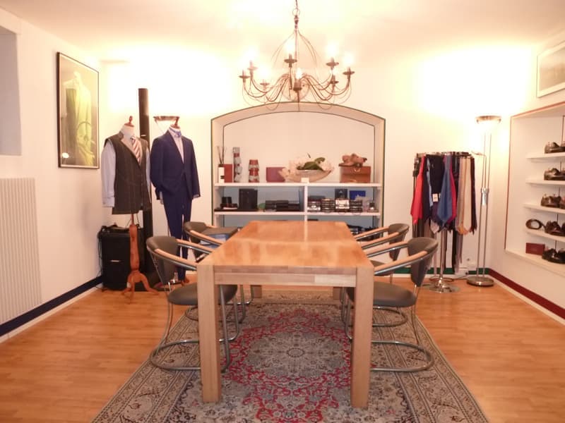A well-lit room with a wooden table and chairs, a chandelier, shelves with various items, and mannequins dressed in suits.