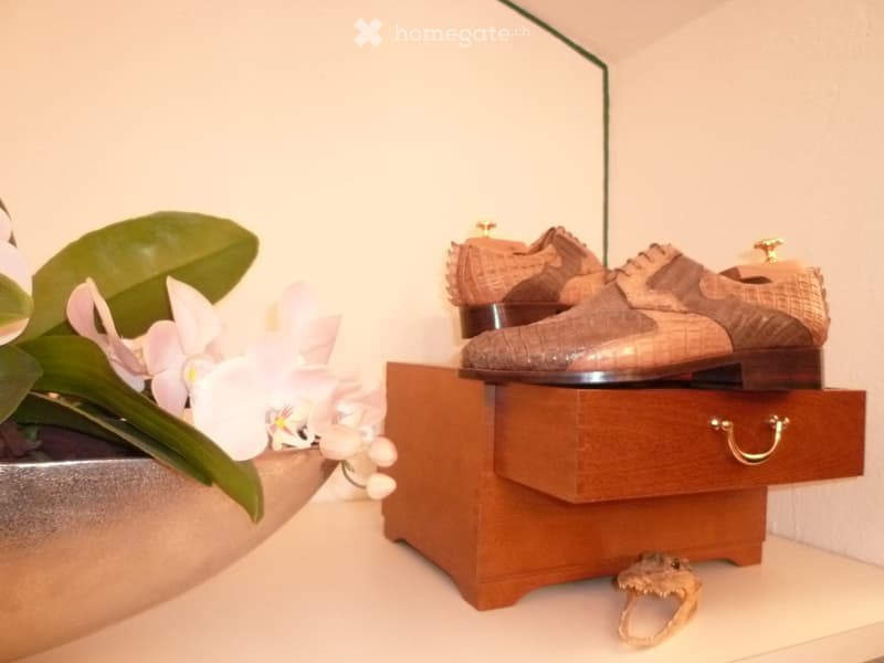 A small wooden cabinet with a drawer, displaying a pair of brown leather shoes and a small flower arrangement in a silver bowl on a white surface.