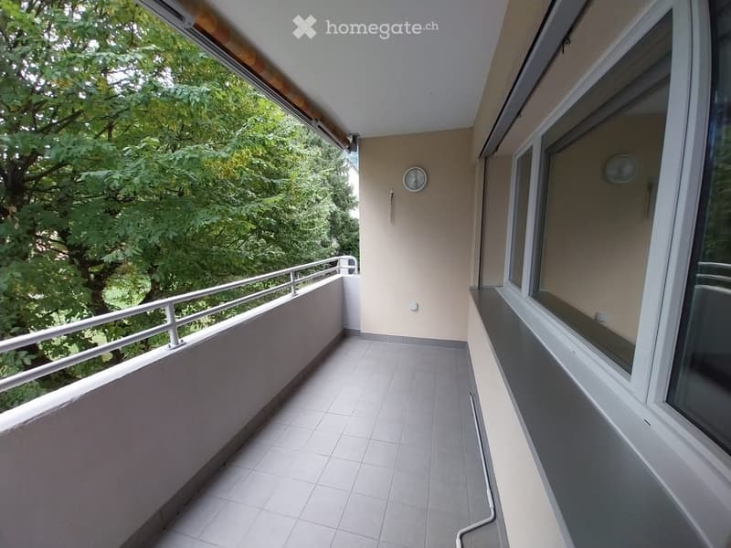 Balcony with tiled floor, metal railing, large windows, and a view of green trees.
