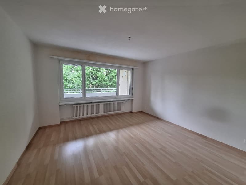 Empty room with wooden flooring, white walls, and large windows with a view of greenery outside.