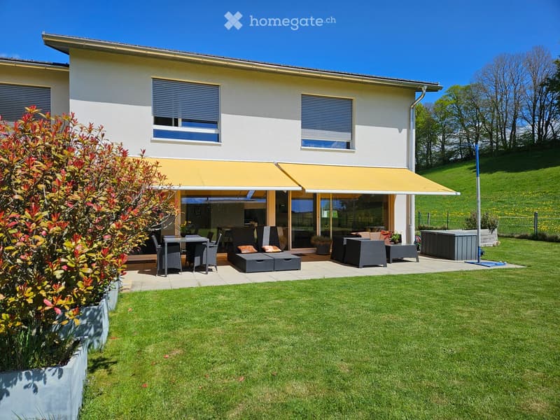 A modern house with a spacious outdoor patio area featuring a dining set, lounge chairs, and a covered seating area. The lawn is well-maintained, and the house has large windows and a yellow awning.
