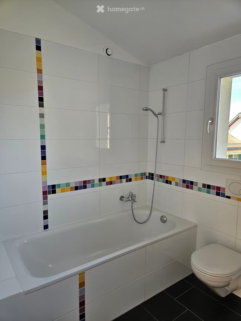 Modern bathroom with a white bathtub, toilet, and colorful mosaic tile accents on the walls. Features a showerhead and faucet above the tub, with a window providing natural light.