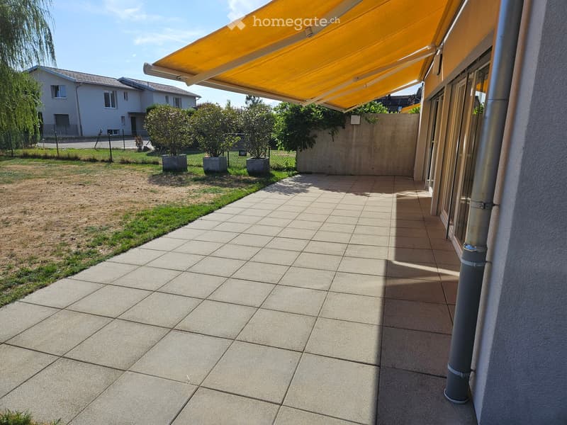 Outdoor patio area with tiled flooring, covered by a yellow awning, surrounded by a low concrete wall, small garden with potted plants, and a grassy area.