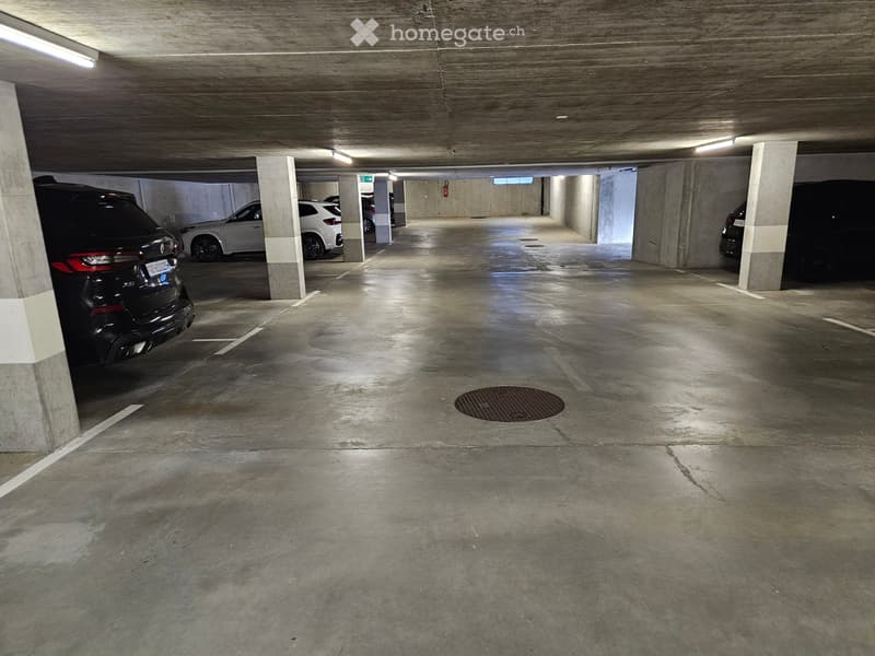 Underground parking area with several parked cars, concrete pillars, and a central drainage grate.