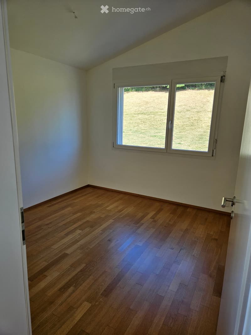 Empty room with wooden flooring, white walls, and a large window overlooking a grassy area.