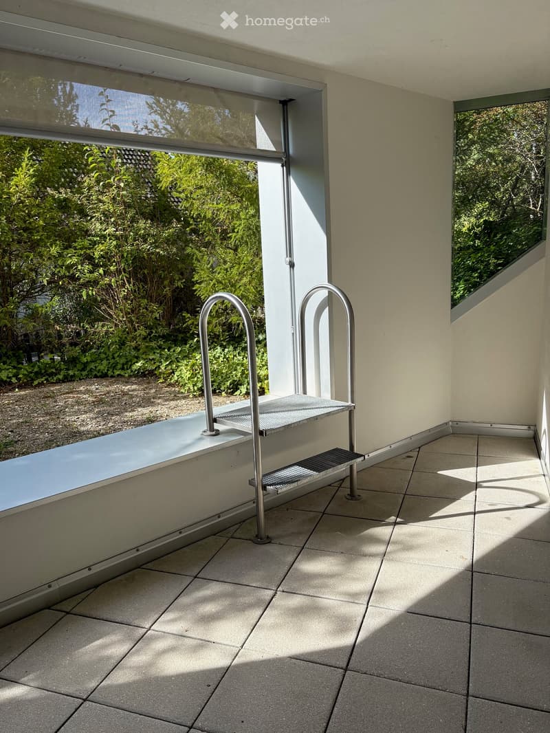 Modern balcony with tiled floor, large glass windows, and metal handrail leading to a lush green garden.