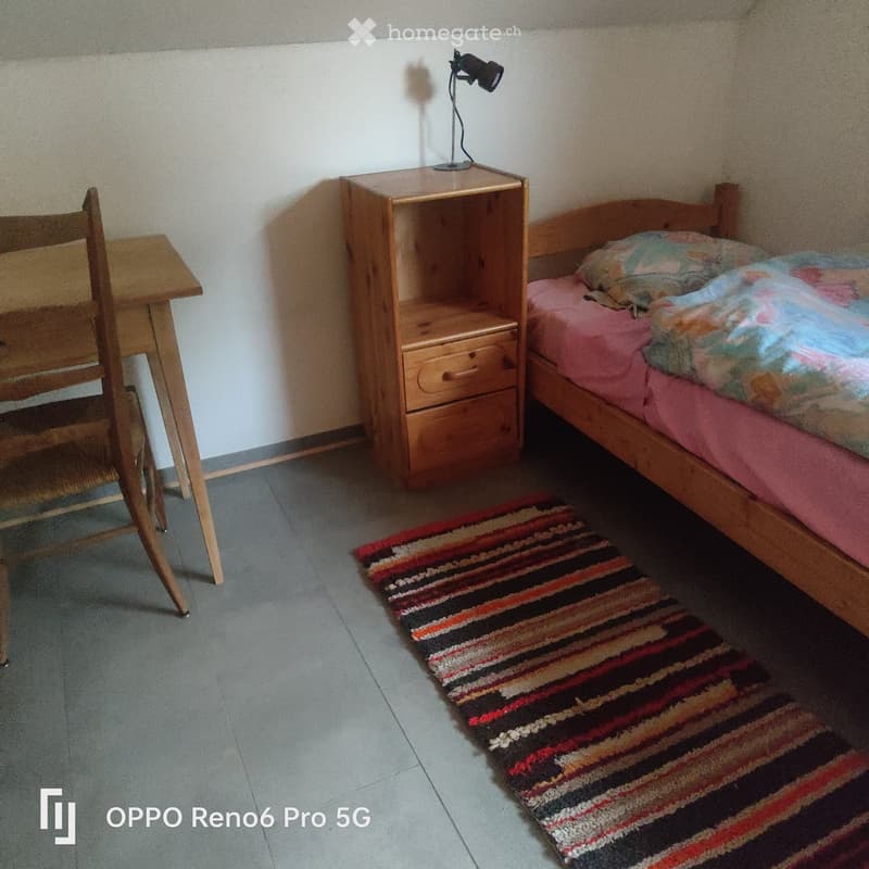 A small bedroom with a wooden bed, nightstand, chair, table, and a colorful striped rug on the floor.