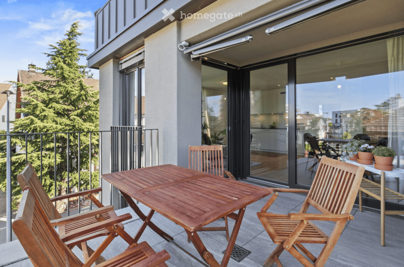 Balcony with wooden table, chairs, and potted plants, overlooking a tree-lined street.