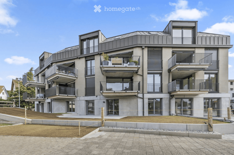 Modern multi-story residential building with balconies, large windows, and a flat roof, surrounded by a paved area and landscaped garden.
