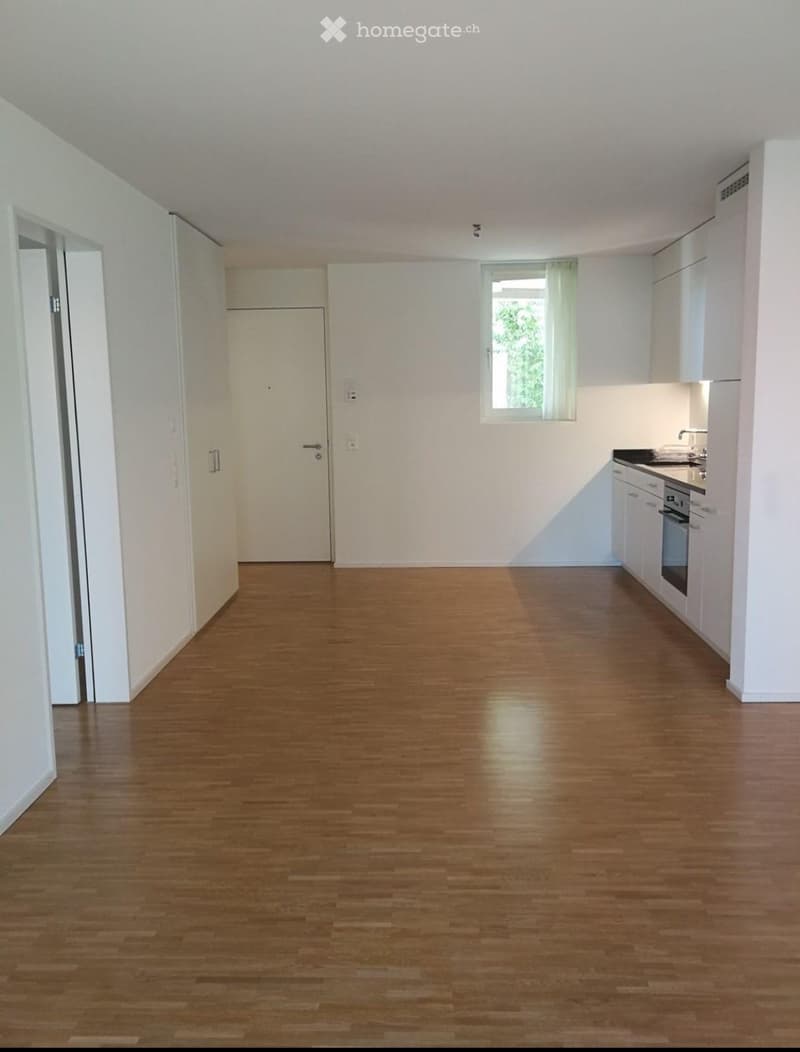 Empty room with wooden flooring, white walls, and a small kitchen area with cabinets and appliances visible in the background.