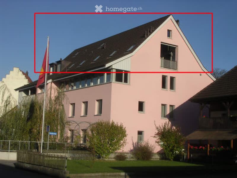 A two-story pink house with white trim, multiple windows, and a balcony on the upper floor. The house has a dark roof and is surrounded by small trees and shrubs.