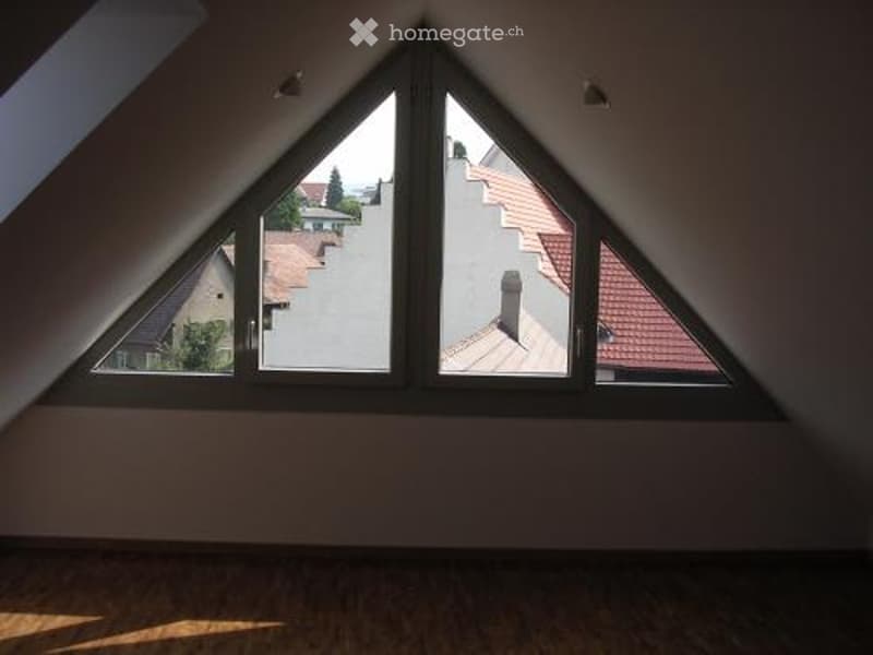 Empty attic room with wooden flooring and large triangular windows offering a view of the neighborhood.