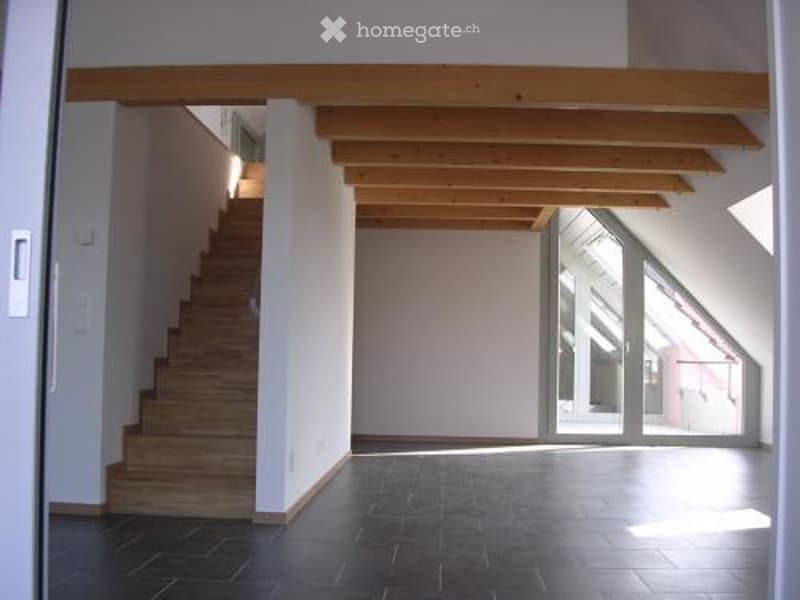 Empty room with wooden staircase, exposed wooden ceiling beams, large window, and dark tiled floor.