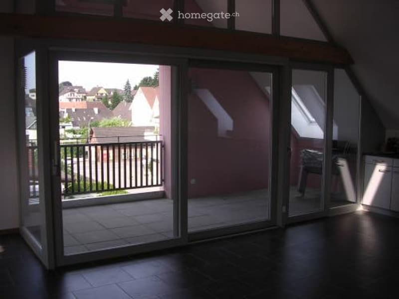 Bright, modern room with large sliding glass doors leading to a balcony. Dark hardwood floor, white cabinetry, and a single chair visible near the balcony.