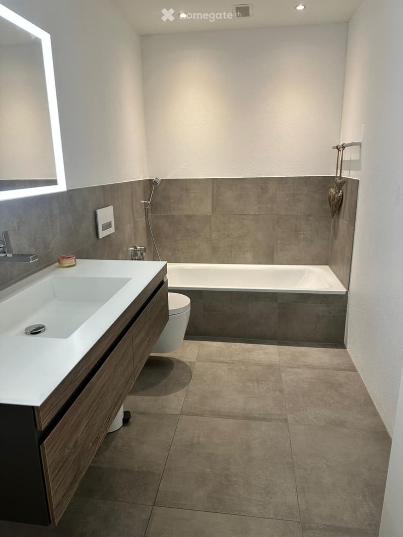 Modern bathroom with a white sink, bathtub, toilet, and a large mirror. The walls are tiled in a neutral color scheme, and the floor is covered with beige tiles.