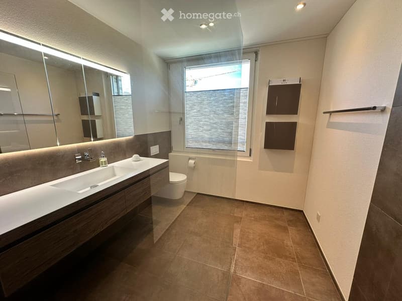 Modern bathroom with a double sink, toilet, large mirror, and built-in cabinets. Features a window with blinds and a towel rack on the wall.