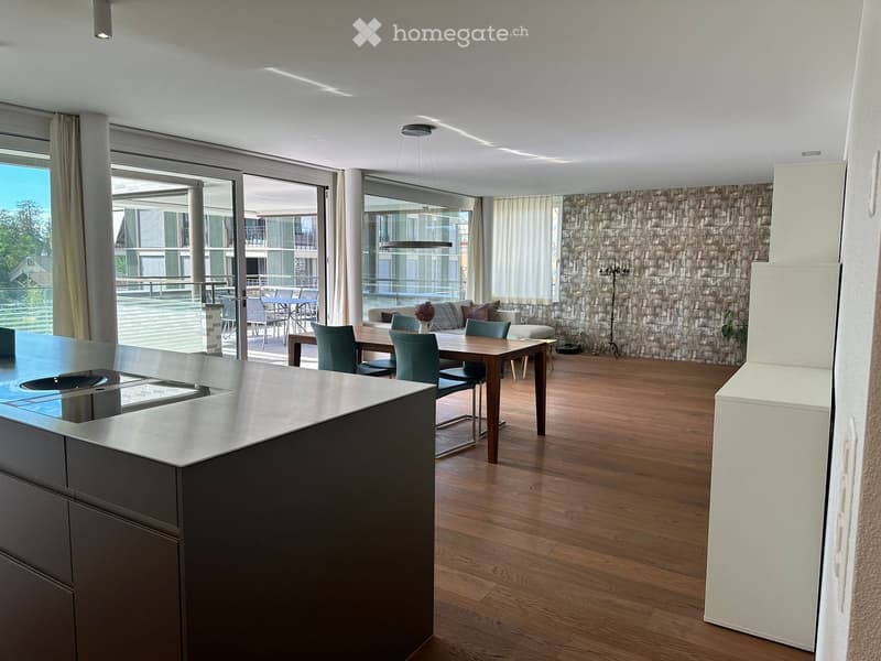 Modern open-plan living and dining area with a kitchen island, dining table, sofa, and ottoman, connected to a balcony with outdoor seating.