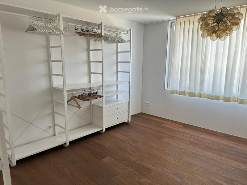 Empty room with wooden flooring, white walls, large window with white curtains, and a white clothing rack with hangers.
