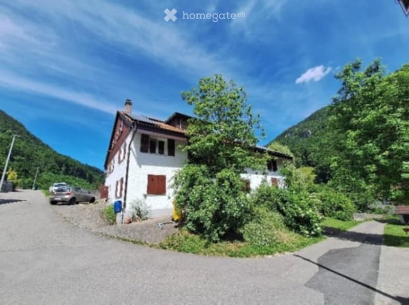 A two-story white house with brown shutters and a tiled roof, situated in a scenic, green valley with mountains in the background. The house has a small driveway with a parked car and is surrounded by lush greenery.