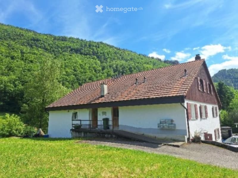 Single-story house with a red-tiled roof, white walls, and a small porch. Surrounded by green grass and trees with a mountain backdrop.