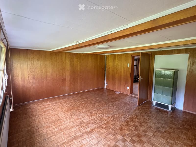 Empty room with wooden walls and herringbone-patterned wooden flooring, featuring a single green cabinet on a stand and a visible doorway leading to another space.