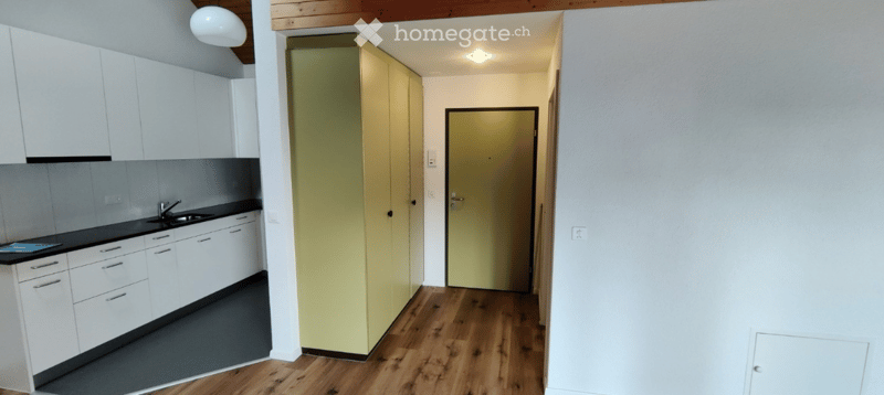 Modern kitchen with white cabinets, black countertops, and a wooden floor. A doorway leads to a hallway with a yellow door.