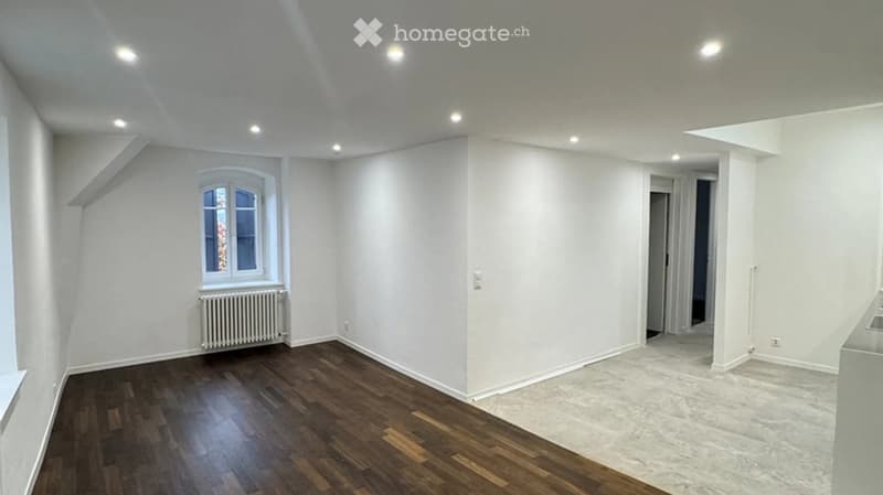Empty room with wooden flooring, white walls, and a single window with a radiator below it. The ceiling has recessed lighting, and there is an open doorway leading to another space.