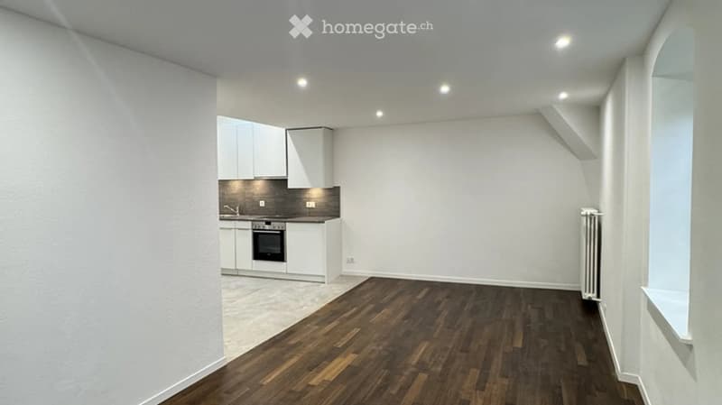 Modern, minimalist room with wooden flooring, white walls, and a compact kitchen area featuring cabinets, an oven, and a faucet.