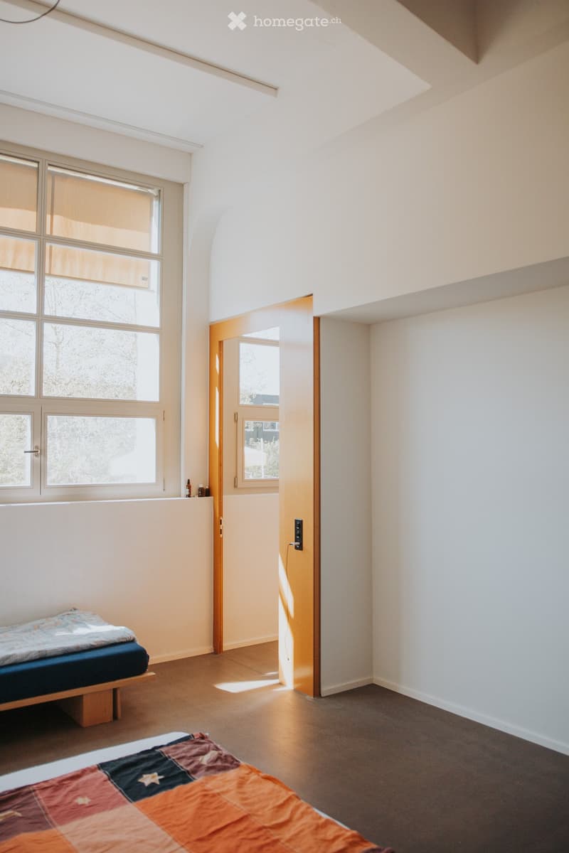 Bright bedroom with two beds, large windows, and a wooden door leading to another room.