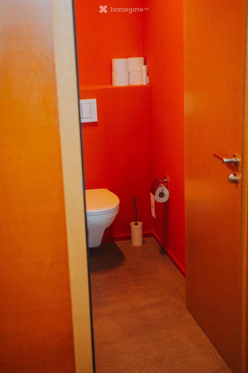 Small bathroom with red walls, white toilet, toilet paper holder, and toilet brush holder. Toilet paper is stored on a shelf above the toilet.