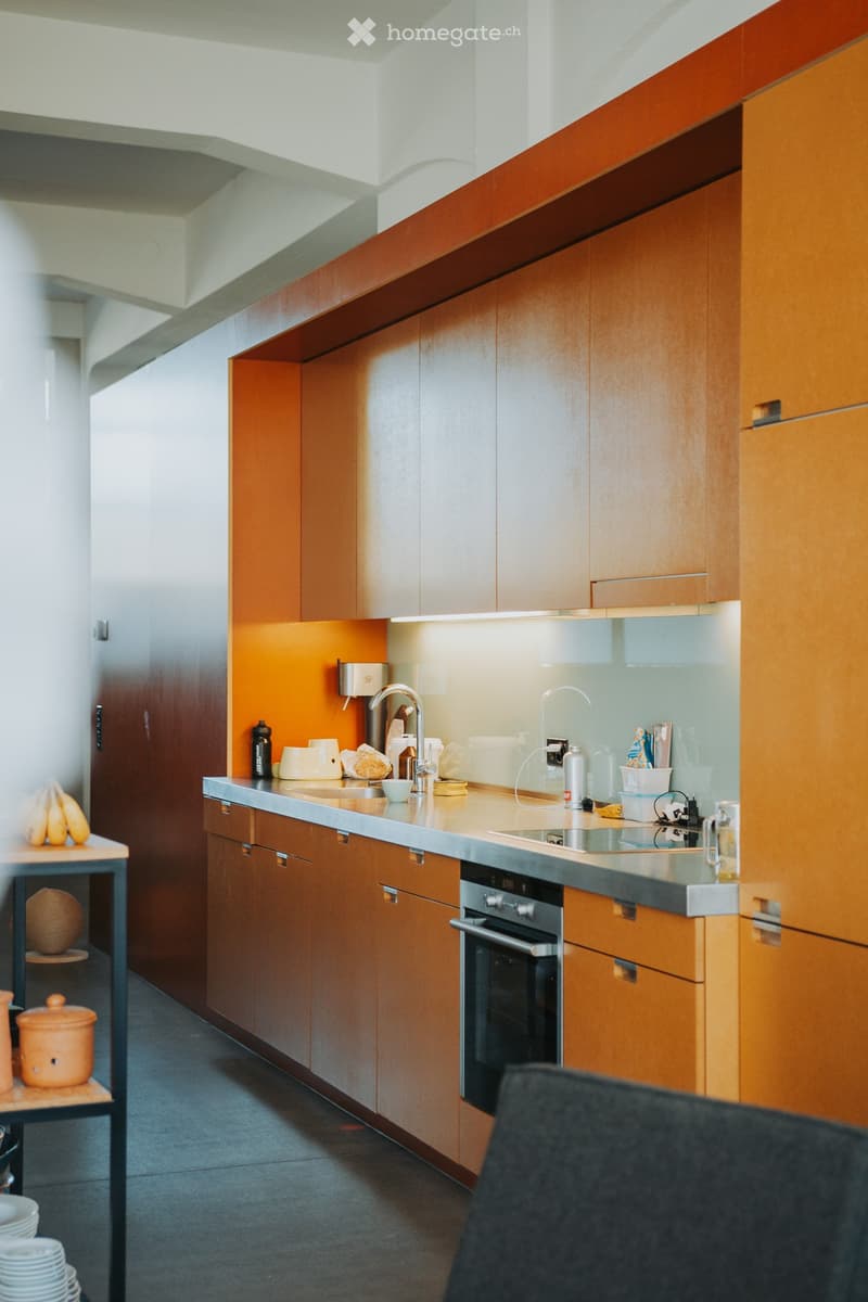 Modern kitchen with wooden cabinets, stainless steel sink, and various appliances. Countertop has a bowl, a water bottle, and other small items. A small table with a ceramic pot and bananas is visible in the foreground.