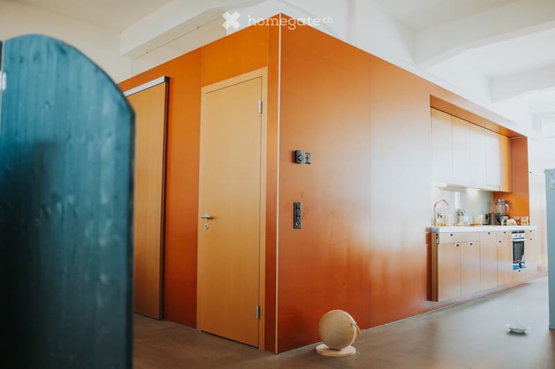 Modern kitchen with light wood cabinets, stainless steel appliances, and a vibrant orange accent wall. Visible items include a faucet, oven, blender, and a decorative ball on the floor.