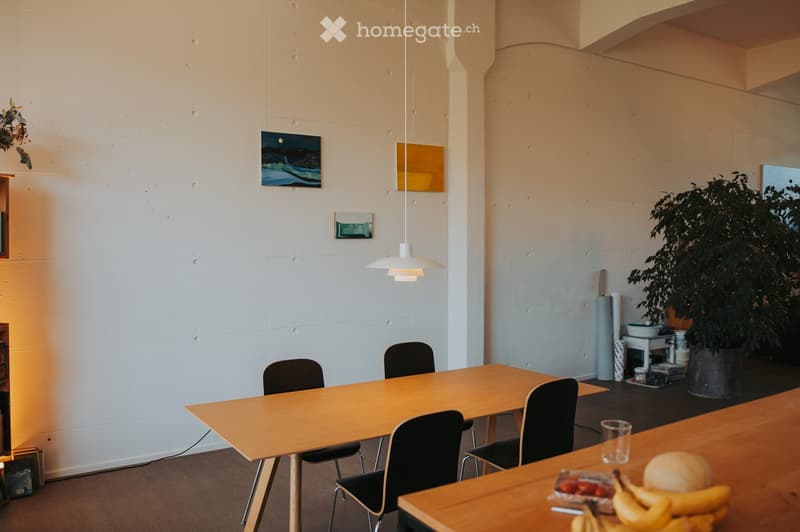 Bright, minimalist room with a wooden dining table surrounded by black chairs, a pendant light hanging above, and a potted plant in the corner. A counter with fruits and a cup is visible.