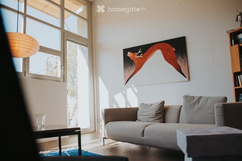 Modern living room with a grey sectional sofa, ottoman, coffee table, pendant light, bookshelf, and a large abstract painting on the wall. Large windows allow natural light to fill the space.