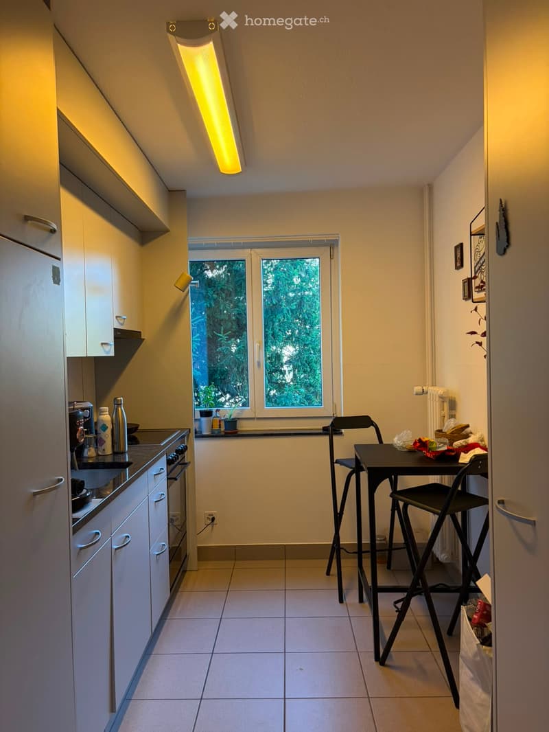 Modern kitchen with white cabinets, stainless steel appliances, a small dining area with a table and chairs, and a window overlooking greenery.