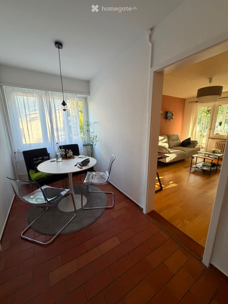 Modern living and dining area with wooden flooring, white walls, and large windows. Features a sofa, coffee table, dining table with chairs, and pendant lighting.